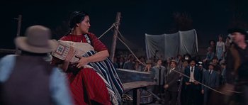 Movie still from “Cimarron” (1960), directed by Charles Walters – A woman sitting on top of a bench in front of a group of people; Medium shot, Low angle
