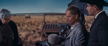 Movie still from “Cimarron” (1960), directed by Charles Walters – A man in a suit and tie holding an umbrella in a field; Medium shot, Low angle