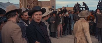 Movie still from “Cimarron” (1960), directed by Charles Walters – A man in a cowboy hat standing in front of a group of people; Medium shot, Over the shoulder angle