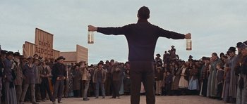Movie still from “Cimarron” (1960), directed by Charles Walters – A man holding up a bottle in front of a group of people; Wide shot, Low angle
