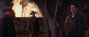 Movie still from “Cimarron” (1960), directed by Charles Walters – A man wearing a hat standing next to a tree; Medium shot, Over the shoulder angle