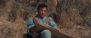 Movie still from “Cimarron” (1960), directed by Charles Walters – A man sitting on the ground holding a wooden board; Medium shot, Low angle