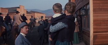 Movie still from “Cimarron” (1960), directed by Charles Walters – A woman holding a child in her arms while standing on a street; Medium shot, Over the shoulder angle