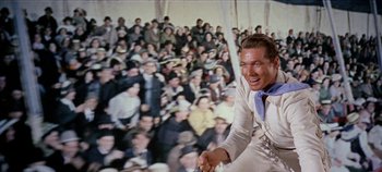 Movie still from “Circus World” (1964), directed by Henry Hathaway – A man in a white suit and blue neck tie smiles in front of a crowd; Medium shot, Low angle