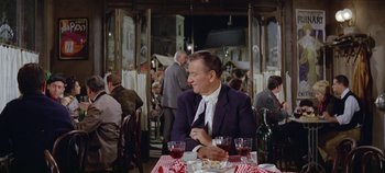 Movie still from “Circus World” (1964), directed by Henry Hathaway – A man sitting at a table with a glass of red wine; Medium shot, Over the shoulder angle