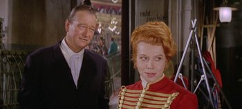 Movie still from “Circus World” (1964), directed by Henry Hathaway – A man standing next to a woman wearing a red dress; Medium shot, Over the shoulder angle