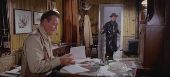 Movie still from “Circus World” (1964), directed by Henry Hathaway – A man in a hat is standing at a table and another man is sitting at a desk; Wide shot, Over the shoulder angle