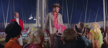 Movie still from “Circus World” (1964), directed by Henry Hathaway – A man wearing a cowboy hat standing in front of a group of people; Medium shot, Over the shoulder angle