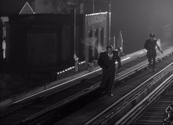 Movie still from “City That Never Sleeps” (1953), directed by John H. Auer – A black and white photo of a man walking on train tracks at night; Wide shot, High angle