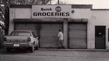 Movie still from “Clerks II” (2006), directed by Kevin Smith – A man standing in front of a store with a car parked in front of it; Wide shot, Low angle