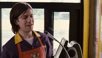 Movie still from “Clerks II” (2006), directed by Kevin Smith – A man in an orange and purple apron standing in front of a microphone; Medium shot, Over the shoulder angle