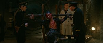 Movie still from “Cliff Walkers” (2021), directed by Yimou Zhang – A man is tied up in a chair with a wooden stick; Medium shot, Over the shoulder angle