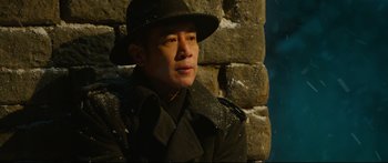 Movie still from “Cliff Walkers” (2021), directed by Yimou Zhang – A man wearing a hat and a trench coat leaning against a stone wall; Close Up shot, Over the shoulder angle