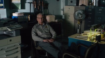 Movie still from “The Dead Don't Die” (2019), directed by Jim Jarmusch – An older man sitting in a chair in front of a desk; Medium shot, Over the shoulder angle