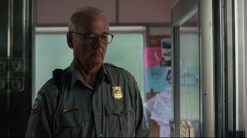 Movie still from “The Dead Don't Die” (2019), directed by Jim Jarmusch – An older man wearing glasses and wearing a police badge; Close Up shot, Over the shoulder angle