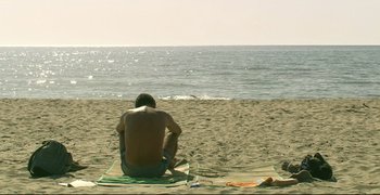 Movie still from “Climates” (2006), directed by Nuri Bilge Ceylan – A man sitting on a towel on the beach looking out at the ocean; Wide shot, High angle