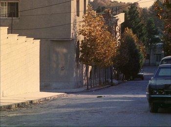 Movie still from “Close-Up” (1990), directed by Abbas Kiarostami – A car parked on the side of the road next to a building; Extreme Wide shot, High angle