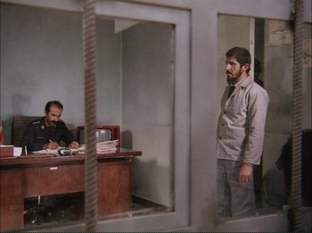 Movie still from “Close-Up” (1990), directed by Abbas Kiarostami – A man standing in front of a man sitting at a desk; Wide shot, Over the shoulder angle