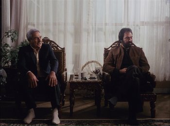 Movie still from “Close-Up” (1990), directed by Abbas Kiarostami – Two men sitting in chairs in front of a window; Wide shot, Low angle
