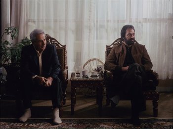 Movie still from “Close-Up” (1990), directed by Abbas Kiarostami – Two men sitting in chairs in a living room next to each other; Wide shot, Over the shoulder angle