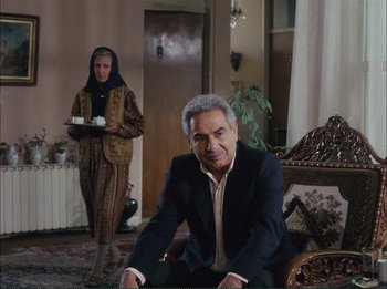 Movie still from “Close-Up” (1990), directed by Abbas Kiarostami – An older man sitting in a chair in front of an older woman; Wide shot, Over the shoulder angle