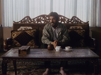 Movie still from “Close-Up” (1990), directed by Abbas Kiarostami – A man sitting at a table with a cup of coffee; Wide shot, Low angle