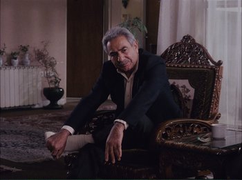 Movie still from “Close-Up” (1990), directed by Abbas Kiarostami – An older man sitting on a chair in a living room; Medium shot, High angle