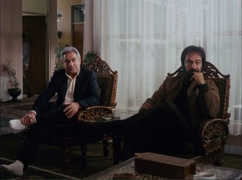 Movie still from “Close-Up” (1990), directed by Abbas Kiarostami – Two men sitting in chairs in a living room next to each other; Wide shot, Low angle