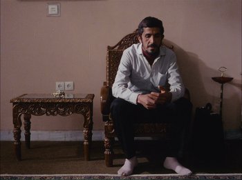 Movie still from “Close-Up” (1990), directed by Abbas Kiarostami – A man sitting on a chair in a room; Medium shot, Low angle