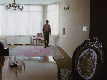 Movie still from “Close-Up” (1990), directed by Abbas Kiarostami – A man standing in a living room next to a rug; Wide shot, High angle