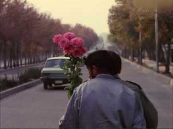 Movie still from “Close-Up” (1990), directed by Abbas Kiarostami – A man walking down the street holding a bouquet of flowers; Medium shot, Over the shoulder angle