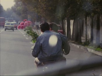Movie still from “Close-Up” (1990), directed by Abbas Kiarostami – A man riding a motorcycle down a street holding flowers; Wide shot, Over the shoulder angle