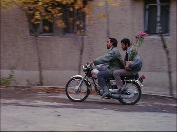 Movie still from “Close-Up” (1990), directed by Abbas Kiarostami – Two men riding a motorcycle with a bouquet of flowers on the back of it; Wide shot, High angle