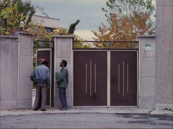 Movie still from “Close-Up” (1990), directed by Abbas Kiarostami – Two men standing in front of an entrance to a building; Wide shot, Over the shoulder angle