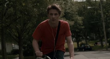 Movie still from “Closet Monster” (2015), directed by Stephen Dunn – A man riding a bike down a street with headphones on; Medium shot, Low angle