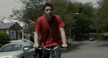 Movie still from “Closet Monster” (2015), directed by Stephen Dunn – A man riding a bike down a street with headphones on; Medium shot, Low angle