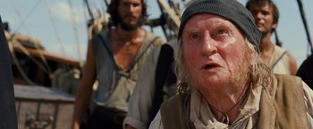 Movie still from “Cloud Atlas” (2012), directed by Lilly Wachowski – A man with a beard and a hat is looking at the camera while another man stands in the background; Close Up shot, Low angle