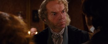 Movie still from “Cloud Atlas” (2012), directed by Lilly Wachowski – A man with long blonde hair wearing a black suit and a white neck tie; Close Up shot, Over the shoulder angle