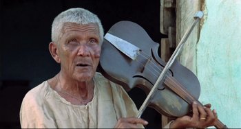 Movie still from “Cobra Verde” (1987), directed by Werner Herzog – An older man holding a violin in front of him; Close Up shot, Low angle