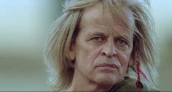 Movie still from “Cobra Verde” (1987), directed by Werner Herzog – An older man with long blonde hair and blue eyes; Close Up shot, Low angle