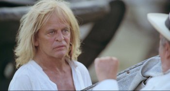 Movie still from “Cobra Verde” (1987), directed by Werner Herzog – A man with long blonde hair wearing a white shirt; Close Up shot, Over the shoulder angle