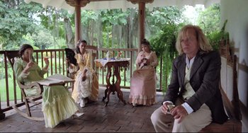 Movie still from “Cobra Verde” (1987), directed by Werner Herzog – A group of people dressed in period clothing sitting on a porch; Wide shot, Over the shoulder angle