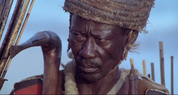 Movie still from “Cobra Verde” (1987), directed by Werner Herzog – An older man with a hat on holding a wooden stick; Close Up shot, Low angle