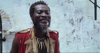 Movie still from “Cobra Verde” (1987), directed by Werner Herzog – A man with a beard wearing a red vest and a necklace; Close Up shot, Low angle