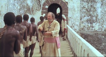 Movie still from “Cobra Verde” (1987), directed by Werner Herzog – An old man holding a cross in front of a group of people; Medium shot, High angle