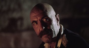 Movie still from “Cobra Verde” (1987), directed by Werner Herzog – A bald man with a mustache and a goatee looking at the camera; Close Up shot, Low angle
