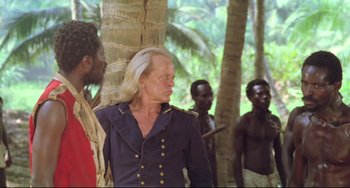 Movie still from “Cobra Verde” (1987), directed by Werner Herzog – A man with long blonde hair standing next to a group of men; Medium shot, Over the shoulder angle