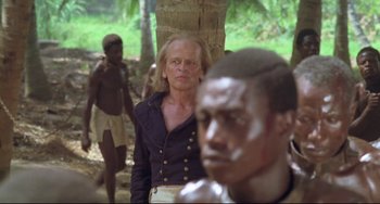 Movie still from “Cobra Verde” (1987), directed by Werner Herzog – A man standing in front of a group of people; Medium shot, Over the shoulder angle