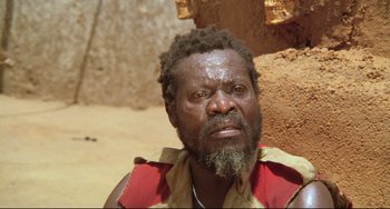 Movie still from “Cobra Verde” (1987), directed by Werner Herzog – A man with a beard and a red shirt is looking at the camera; Close Up shot, Low angle