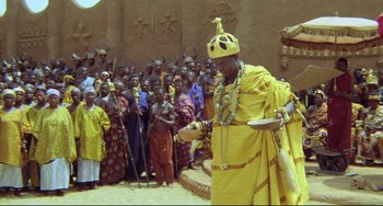 Movie still from “Cobra Verde” (1987), directed by Werner Herzog – A man in a yellow robe is holding a bowl; Wide shot, Low angle